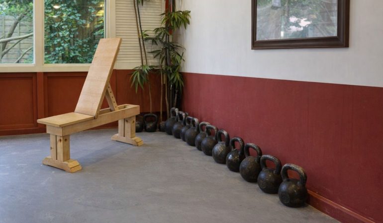En träningsbänk i trä och en rad kettlebells längs en vägg.