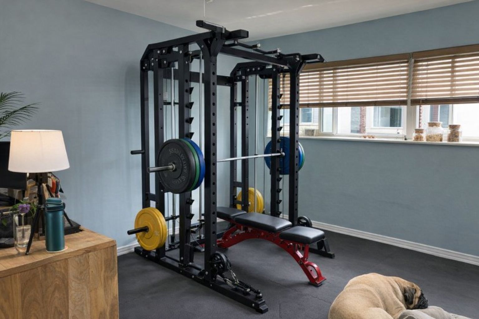 En robust svart smithmaskin med skivstång och träningsbänk i ett hemgym.